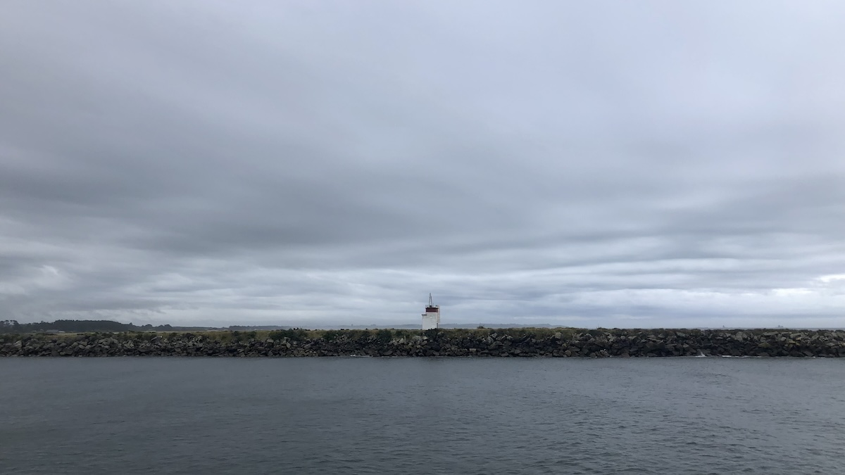 Photo of the harbour mouth in Westport. Grey sea in the lower third, then a strip of land with a small building, and above a foreboding grey sky.